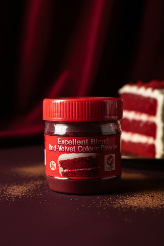 A hand holding a clear plastic jar of red velvet cake mix powder with a red lid and red label. The label reads 'Excellent Blend Red-Velvet Colour Powder' and shows a slice of red velvet cake.