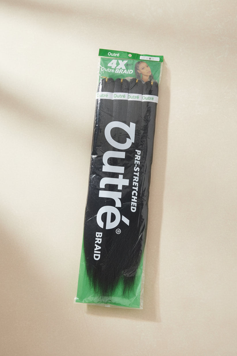 A package of outre 4X Dutre Braid Pre-Stretched black hair extensions laid flat on a dark wood floor. The packaging is clear plastic with green and white branding.